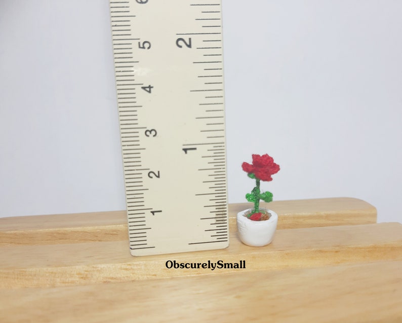 May include: A miniature crocheted red rose in a white ceramic pot. The green stem and leaves support the flower. The pot is approximately 1 inch tall. The text "ObscurelySmall" is visible.
