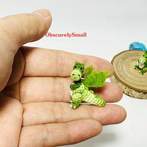 May include: Two small green crocheted dragons with white accents. One dragon is being held in a hand, while the other is sitting on a wooden surface.