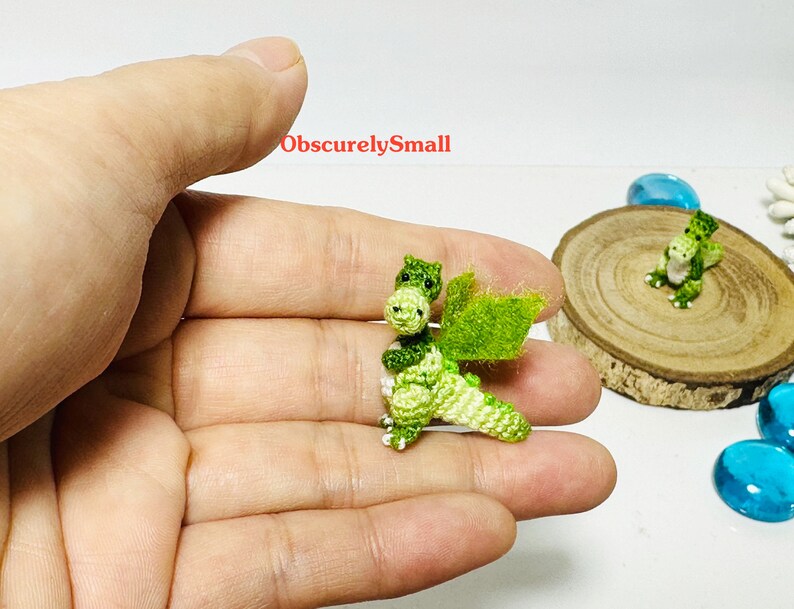 May include: Two small green crocheted dragons with white accents. One dragon is being held in a hand, while the other is sitting on a wooden surface.