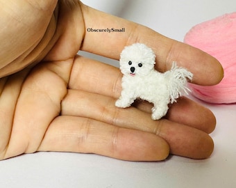 Może przedstawiać: Miniaturowa figurka białego psa o puszystej fakturze, z czarnymi oczami i czarnym nosem. Nad psem znajduje się napis ObscurelySmall. Figurka wykonana jest z drobnych włókien przypominających rasę Bichon Frise i posiada mały ogonek.
