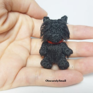 May include: A small black crocheted dog with a red collar. The dog is sitting on a hand.