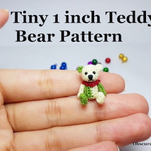 Peut inclure: Un petit ours en peluche crocheté, d'environ 2,5 cm de haut, est assis sur une main. L'ours est blanc avec des vêtements verts et un collier rouge. Le texte "Tiny 1 inch Teddy Bear Pattern" est en haut de l'image.