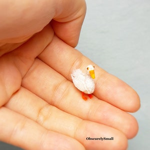 May include: A tiny white crocheted goose with orange feet and a yellow beak. The goose is sitting in the palm of a hand.