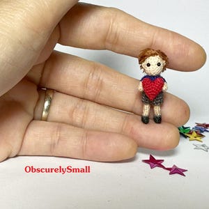May include: A miniature crocheted doll of a boy with brown hair, a red heart, and gray shorts. The doll is held in a hand, with the text "ObscurelySmall" visible. Colorful star-shaped confetti is scattered nearby.