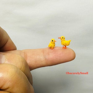 May include: Two tiny crocheted yellow chicks, one standing upright and the other perched on a finger, with orange feet.