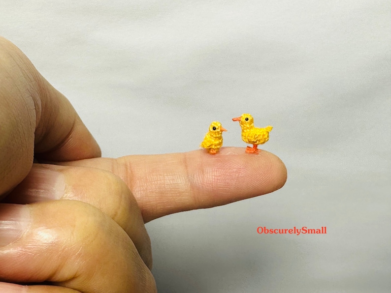 May include: Two tiny crocheted yellow chicks, one standing upright and the other perched on a finger, with orange feet.