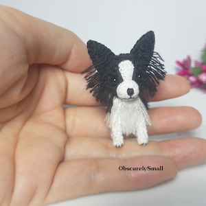 May include: A small crocheted black and white dog with long fur. The dog is sitting on a hand.