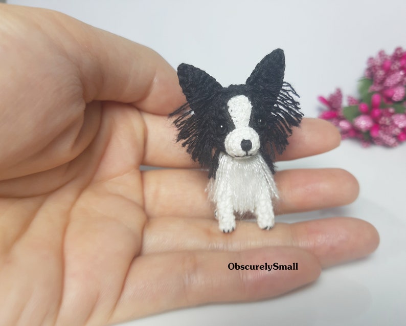May include: A small crocheted black and white dog with long fur. The dog is sitting on a hand.