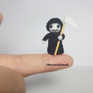 May include: A miniature crocheted Grim Reaper figure, dressed in a black hooded robe, holding a white scythe. The figure has a skull-like face and is standing on a fingertip. The text "ObscurelySmall" is visible.