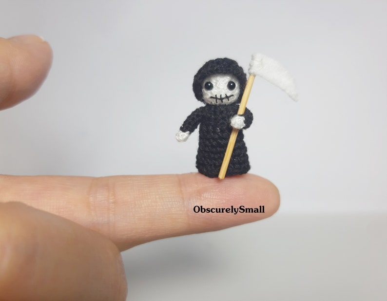 May include: A miniature crocheted Grim Reaper figure, dressed in a black hooded robe, holding a white scythe. The figure has a skull-like face and is standing on a fingertip. The text "ObscurelySmall" is visible.