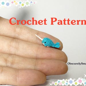 May include: A tiny, turquoise crochet narwhal with a white tusk rests on a hand. The image includes the text "Crochet Pattern" in red, and the seller's name, "ObscurelySmall". The narwhal is a miniature handmade craft.