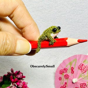 May include: A small green crocheted frog sitting on a red pencil. The frog has black eyes and a white belly.