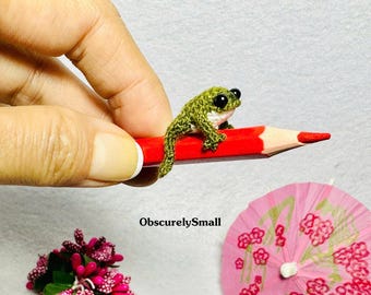 Op de afbeelding: Een kleine groene gehaakte kikker die op een rood potlood zit. De kikker heeft zwarte ogen en een witte buik.