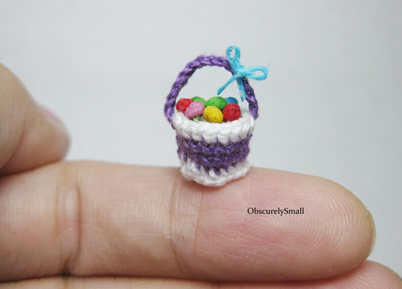 Pu&ograve; includere: Un piccolo cestino all'uncinetto viola e bianco pieno di uova di Pasqua colorate. Il cestino ha un nastro blu legato al manico. Il cestino &egrave; appoggiato su un dito.