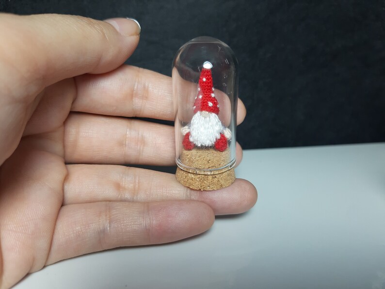 May include: A small, red and white gnome with a white beard is inside a clear glass jar with a cork stopper. The gnome is sitting on a small bed of brown material.
