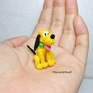 May include: A small crocheted plush toy of Pluto, a yellow dog with black ears, a green collar, and a pink tongue. The toy is sitting on a person's hand.