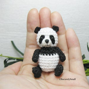 Puede incluir: Un pequeño oso panda de crochet con pelaje blanco y negro. El panda está sentado en una mano.
