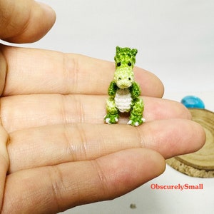 May include: A small green and white crocheted dinosaur toy. The dinosaur is sitting on a person's hand.