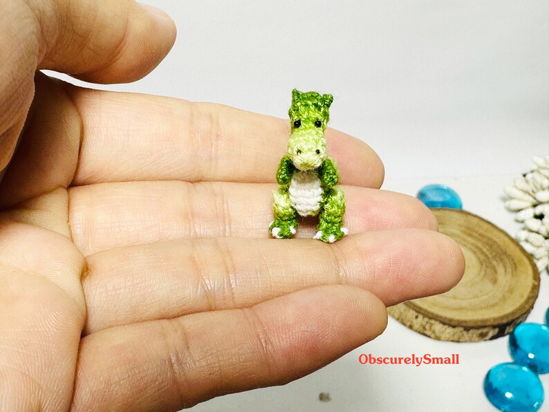 May include: A small green and white crocheted dinosaur toy. The dinosaur is sitting on a person's hand.
