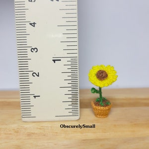 May include: A miniature crocheted sunflower in a woven basket. The sunflower has bright yellow petals and a brown center. The green stem and leaves are visible. The basket is approximately 1 inch tall.