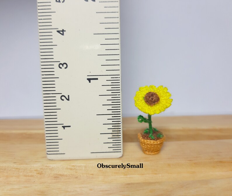 May include: A miniature crocheted sunflower in a woven basket. The sunflower has bright yellow petals and a brown center. The green stem and leaves are visible. The basket is approximately 1 inch tall.