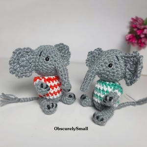 May include: Two small gray crocheted elephant figurines sitting side by side. One elephant wears a red and white striped sweater, while the other wears a green and white striped sweater. Both have black eyes and tails. The text ObscurelySmall is visible.