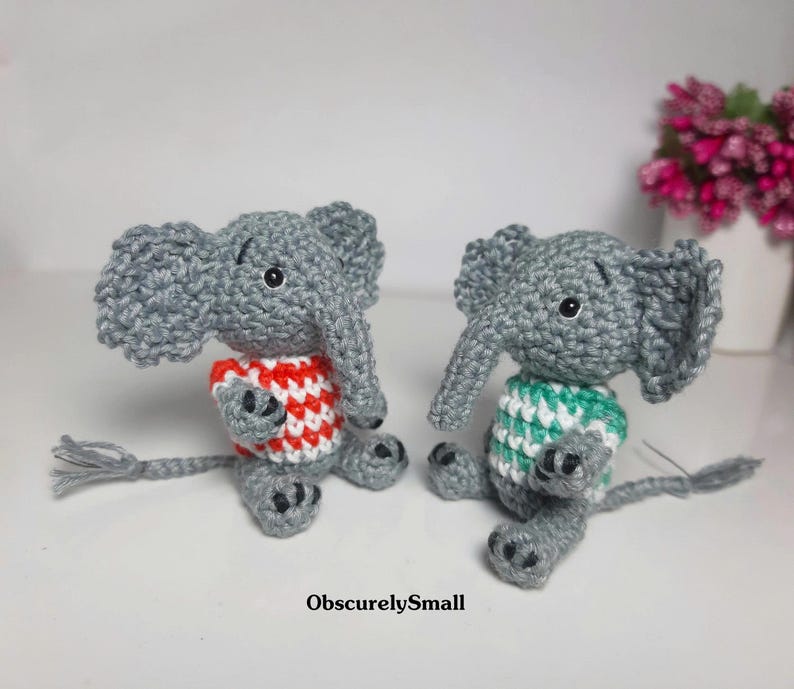 May include: Two small gray crocheted elephant figurines sitting side by side. One elephant wears a red and white striped sweater, while the other wears a green and white striped sweater. Both have black eyes and tails. The text ObscurelySmall is visible.