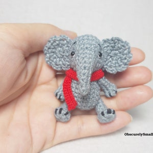 May include: A small, gray crocheted elephant figurine featuring large ears, a trunk, and a bright red scarf wrapped around its neck. The elephant has black bead eyes and small black stitched details on its feet. The text ObscurelySmall is visible.