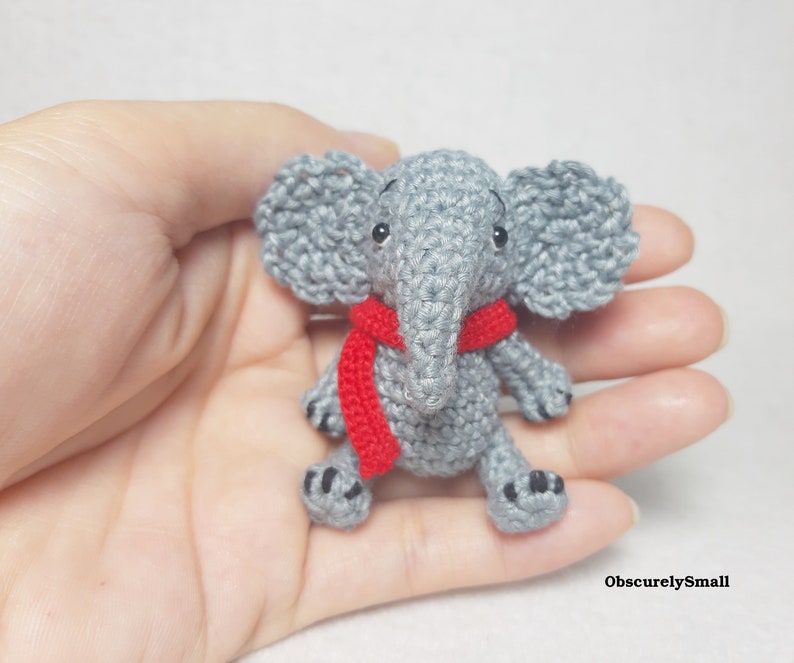 May include: A small, gray crocheted elephant figurine featuring large ears, a trunk, and a bright red scarf wrapped around its neck. The elephant has black bead eyes and small black stitched details on its feet. The text ObscurelySmall is visible.