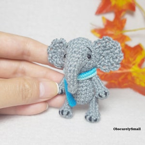 May include: A small gray crocheted elephant figurine featuring large ears, a trunk, and a bright blue scarf wrapped around its neck. The elephant has black embroidered eyes and small black details on its feet. The text ObscurelySmall is visible.