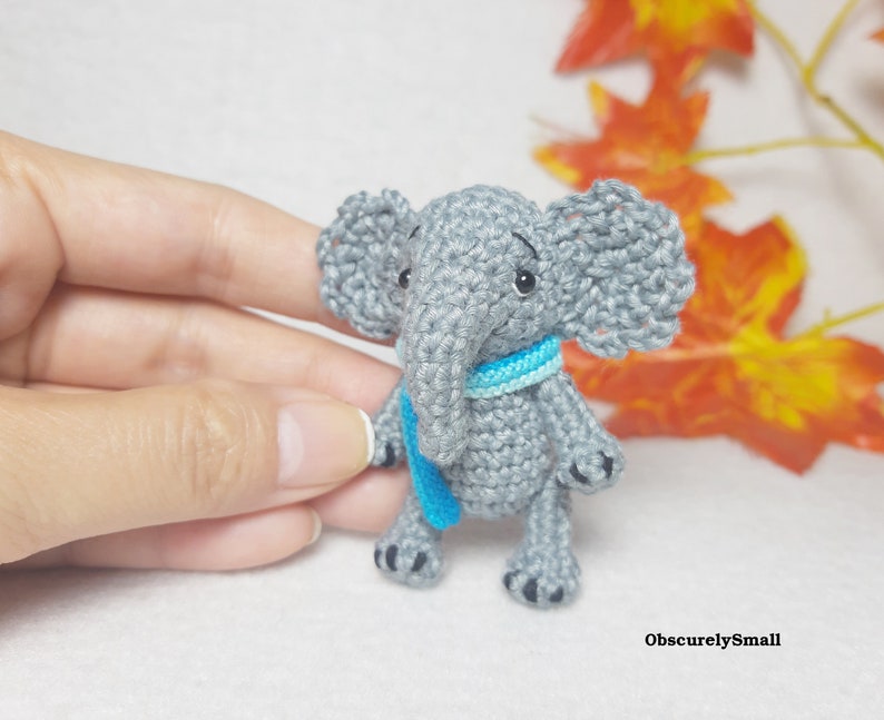 May include: A small gray crocheted elephant figurine featuring large ears, a trunk, and a bright blue scarf wrapped around its neck. The elephant has black embroidered eyes and small black details on its feet. The text ObscurelySmall is visible.