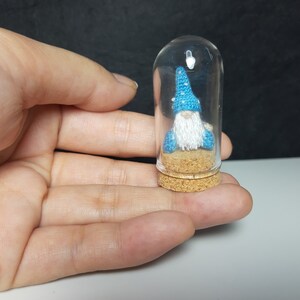 May include: A small blue gnome with a white beard is sitting inside a clear glass jar with a cork lid. The gnome is wearing a blue hat with white stars.