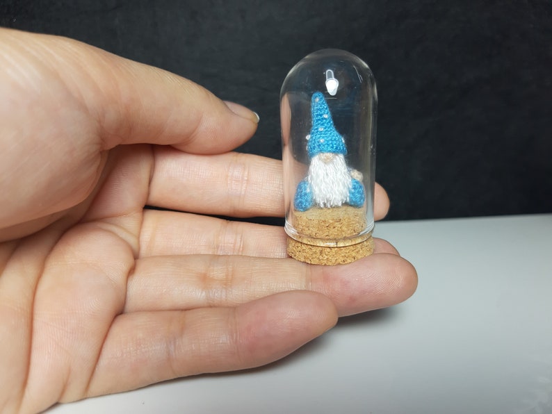 May include: A small blue gnome with a white beard is sitting inside a clear glass jar with a cork lid. The gnome is wearing a blue hat with white stars.
