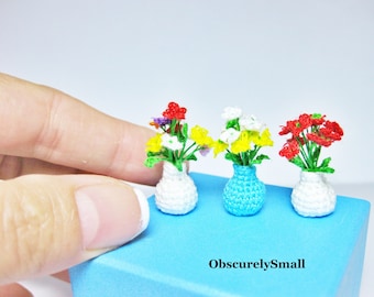 Puede incluir: Tres arreglos florales en miniatura de ganchillo en jarrones blancos, azules y blancos. Las flores son rojas, amarillas y blancas. Los jarrones están sobre una superficie azul. ObscurelySmall está escrito en la superficie.