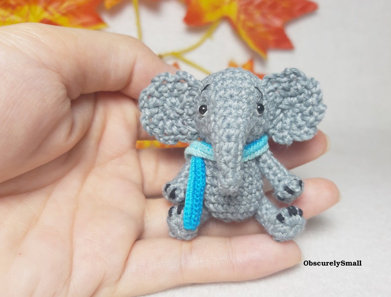 May include: A small, grey crocheted elephant figurine featuring large, textured ears, black bead eyes, and a bright blue scarf wrapped around its neck. The elephant has black stitched details on its feet and the text ObscurelySmall in the corner.