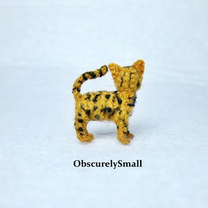 Pu&ograve; includere: Una piccola figurina di gatto all'uncinetto con pelliccia nera e gialla. Il gatto &egrave; rivolto verso la telecamera e ha la coda arrotolata attorno al suo corpo.