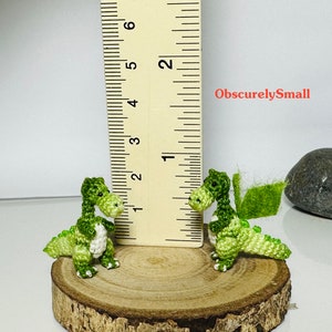 May include: Two small green crocheted dragons, one with wings, are sitting on a wooden log. A ruler is in the background, showing the dragons are about 1 inch tall. The text "ObscurelySmall" is visible in the image.