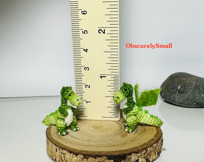 May include: Two small green crocheted dragons, one with wings, are sitting on a wooden log. A ruler is in the background, showing the dragons are about 1 inch tall. The text "ObscurelySmall" is visible in the image.