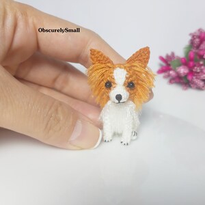 May include: A small crocheted Papillon dog with white fur and orange ears. The dog is sitting on a white surface.
