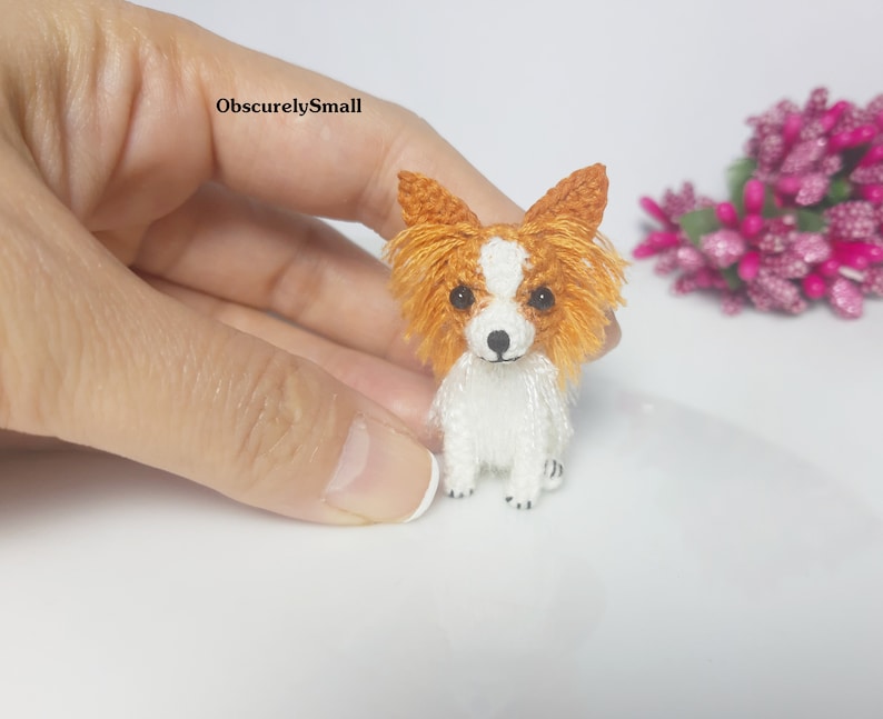 May include: A small crocheted Papillon dog with white fur and orange ears. The dog is sitting on a white surface.
