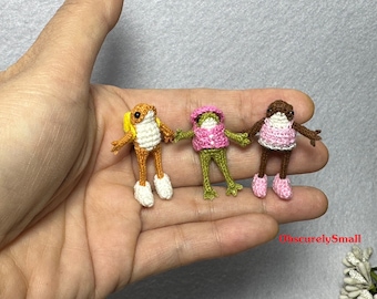 Puede incluir: Tres peluches de rana de crochet, cada una con ropa de diferentes colores. La rana de la izquierda lleva ropa naranja y amarilla, la rana del medio lleva ropa verde y rosa, y la rana de la derecha lleva ropa marrón y rosa.