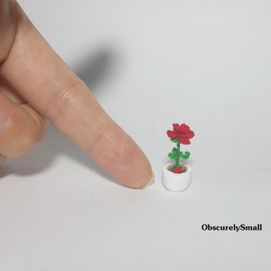 May include: A miniature red rose in a white pot. The rose has green leaves and stem. The pot is small and round. The image is of a handmade, miniature craft item. The text "ObscurelySmall" is in the bottom right corner.