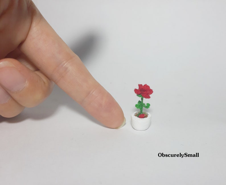 May include: A miniature red rose in a white pot. The rose has green leaves and stem. The pot is small and round. The image is of a handmade, miniature craft item. The text "ObscurelySmall" is in the bottom right corner.