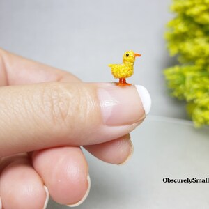 May include: A tiny crocheted yellow duck with orange feet and beak. The duck is sitting on a finger.