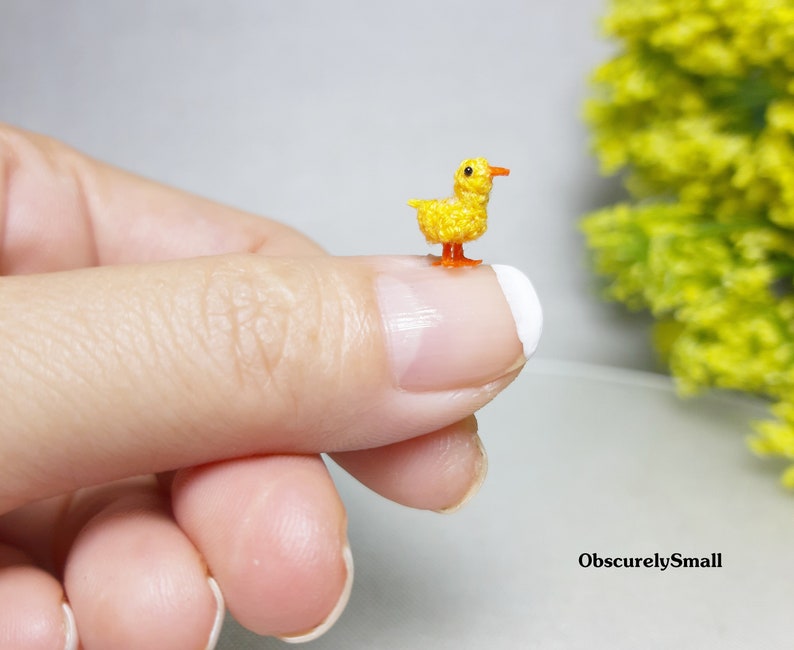 May include: A tiny crocheted yellow duck with orange feet and beak. The duck is sitting on a finger.