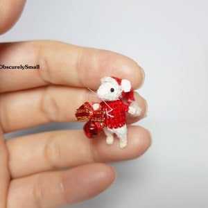May include: A small crocheted mouse wearing a red and white Santa hat and holding a red and gold gift. The mouse is white with black eyes and a pink nose.