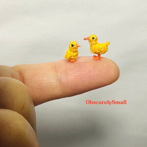 May include: Two tiny crocheted yellow ducklings, one slightly larger than the other, are perched on a finger. The ducklings have orange beaks and feet.