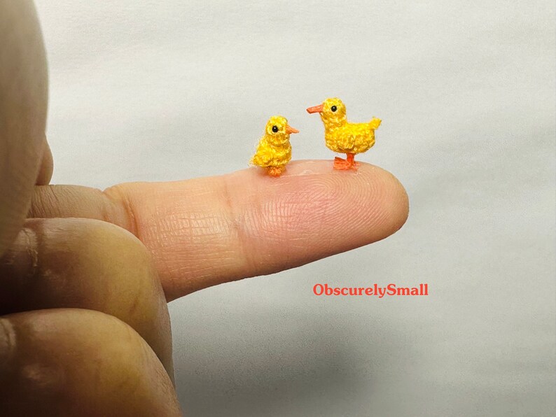 May include: Two tiny crocheted yellow ducklings, one slightly larger than the other, are perched on a finger. The ducklings have orange beaks and feet.