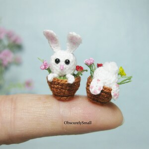 May include: Two white crocheted bunnies in brown pots with pink and red flowers. The bunnies are sitting on a finger.