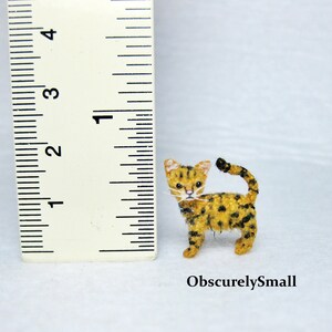 Pu&ograve; includere: Una piccola figurina di gatto in feltro fatta a mano con un motivo a macchie nere e gialle. Il gatto &egrave; in piedi su una superficie bianca accanto a un righello, misurando circa 2,5 cm di altezza.